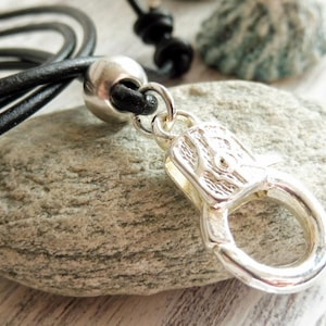 Może przedstawiać: Czarny skórzany naszyjnik ze srebrnym zapięciem i srebrną koralikiem. Naszyjnik leży na szarym kamieniu.