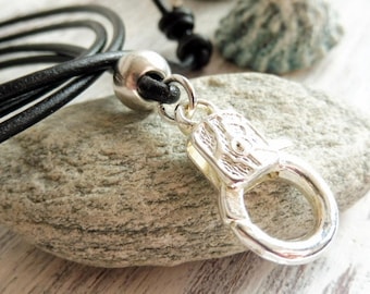 Peut inclure: Un collier en cordon de cuir noir avec un fermoir argenté et une perle argentée. Le collier est posé sur une roche grise.