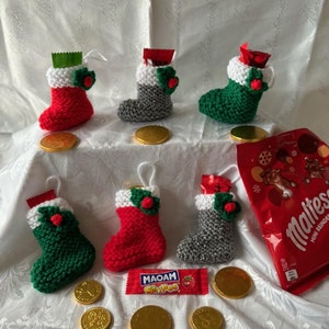 May include: Six small knitted Christmas stockings filled with sweets and chocolate coins. The stockings are red, green, and grey with white trim. The sweets include Maltesers, chocolate coins, and a chocolate bar.