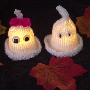 May include: Two white knitted ghost ornaments with googly eyes. The ghost on the left has a pink bow. Both ghosts are lit from within, creating a warm glow.