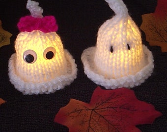 May include: Two white knitted ghost ornaments with googly eyes. The ghost on the left has a pink bow. Both ghosts are lit from within, creating a warm glow.
