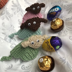 May include: Three knitted Yoda-like figures in pink, brown, and green, with large eyes, sitting on a white background with chocolate eggs and wrappers.