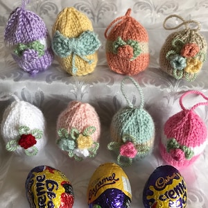 Peut inclure: Dix œufs de Pâques tricotés dans différentes couleurs, dont le violet, le jaune, l'orange, le blanc, le rose et le vert, avec des embellissements floraux et des nœuds. Trois œufs en chocolat enveloppés de papier d'aluminium sont également inclus.