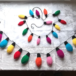 May include: A colourful knitted garland of light bulbs, strung together with black thread. The garland is made up of various colours, including red, orange, yellow, green, blue, and pink.