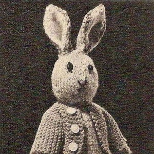 May include: A knitted gray bunny rabbit wearing a gray knitted jacket with white buttons. The bunny is standing on a light gray surface.