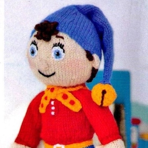 Op de afbeelding: Handgemaakte gebreide pop met een blauwe muts, rood shirt en blauwe korte broek. De pop heeft een gele kraag, een gele riem en rode schoenen. De pop heeft een lachend gezicht en is een kinderspeelgoed.