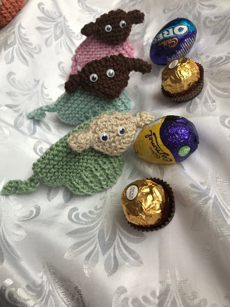May include: Three knitted Yoda-like figures in pink, brown, and green, with large eyes, sitting on a white background with chocolate eggs and wrappers.