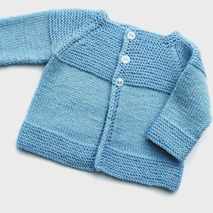 May include: A light blue knitted baby cardigan with three white buttons down the front. The cardigan features a ribbed hem and cuffs, and a contrasting horizontal band across the chest. The soft, textured knitwear is a classic style.