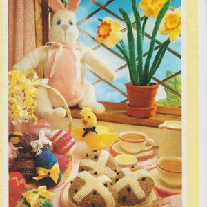 May include: A knitted Easter scene with a white bunny, yellow chick, and a basket of colorful eggs. There are also knitted hot cross buns, a potted plant with yellow daffodils, and a tea set.