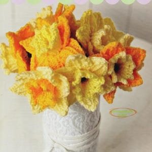 May include: A display of crocheted daffodils in yellow and orange hues. The flowers are presented in a white vase with a patterned design and a white ribbon. The theme is spring and handmade crafts, with a focus on the floral arrangement.