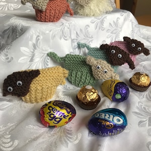 May include: A collection of knitted sheep toys with googly eyes, arranged on a white tablecloth with chocolate eggs. The eggs are wrapped in foil and have the brand names Cadbury Creme Egg and Cadbury Oreo.