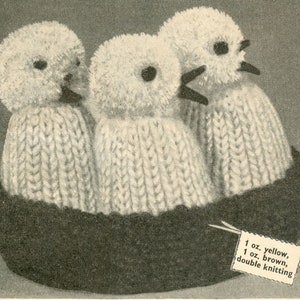 May include: Three knitted white birds with black eyes and beaks are sitting in a black nest. The yarn label reads "1 oz. yellow, 1 oz. brown, double knitting".
