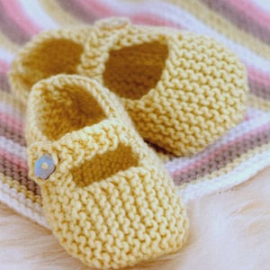 May include: A pair of soft, yellow knitted baby shoes with a Mary Jane style. The shoes have a strap with a button closure and are placed on a striped blanket. The knitted texture is visible.