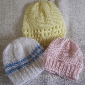 Pode incluir: Três chapéus de tricô para bebê em diferentes cores: amarelo, branco com listras azuis e rosa. Os chapéus são feitos com um padrão de ponto simples e têm uma forma clássica de gorro.