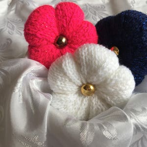 May include: Three knitted flower decorations in pink, white, and blue with gold centers. The flowers are arranged on a white fabric background.