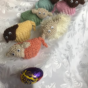 May include: Seven knitted lambs in different colors, including brown, pink, green, beige, and white, with googly eyes. A purple and gold foil-wrapped Cadbury Creme Egg is in the foreground.