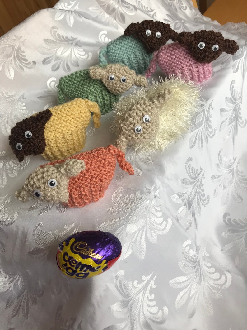 May include: Seven knitted lambs in different colors, including brown, pink, green, beige, and white, with googly eyes. A purple and gold foil-wrapped Cadbury Creme Egg is in the foreground.