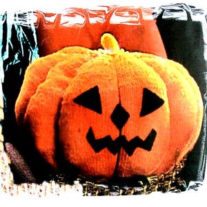 May include: A plush orange pumpkin with a carved jack-o'-lantern face. The pumpkin has black triangle eyes, a diamond-shaped nose, and a jagged mouth. A small yellow stem sits atop the pumpkin. The background is dark.