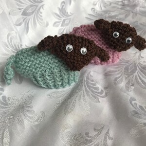 May include: Two crocheted stuffed animals with brown heads and large eyes. One is wearing a light green hat and the other is wearing a light pink hat.
