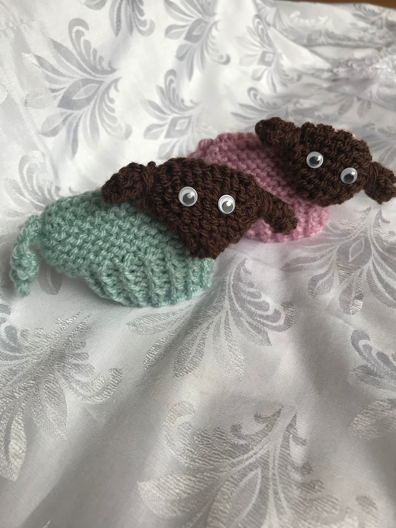 May include: Two crocheted stuffed animals with brown heads and large eyes. One is wearing a light green hat and the other is wearing a light pink hat.