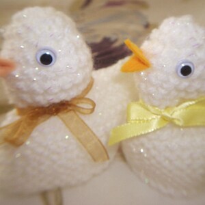 May include: Two white crocheted chicks with yellow beaks and black eyes. One chick has a gold ribbon around its neck and the other has a yellow ribbon.