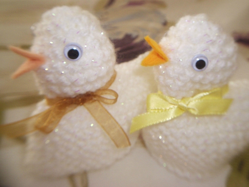 May include: Two white crocheted chicks with yellow beaks and black eyes. One chick has a gold ribbon around its neck and the other has a yellow ribbon.