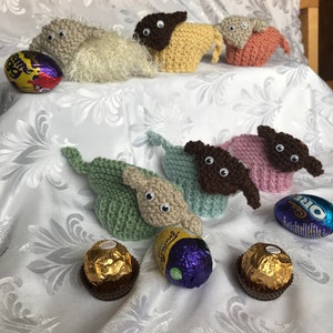 May include: A collection of knitted animal figurines, including a brown and white sheep, a yellow sheep, a pink sheep, a green sheep, a brown sheep, and a pink sheep. The figurines are sitting on a white tablecloth with chocolate eggs and candy.