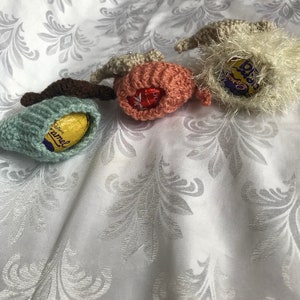 May include: Three crocheted Easter egg covers, one in light blue, one in pink, and one in white. Each cover has a chocolate egg inside, with the brand names "Cadbury Caramel", "Cadbury Dairy Milk", and "Cadbury Creme Egg" visible.