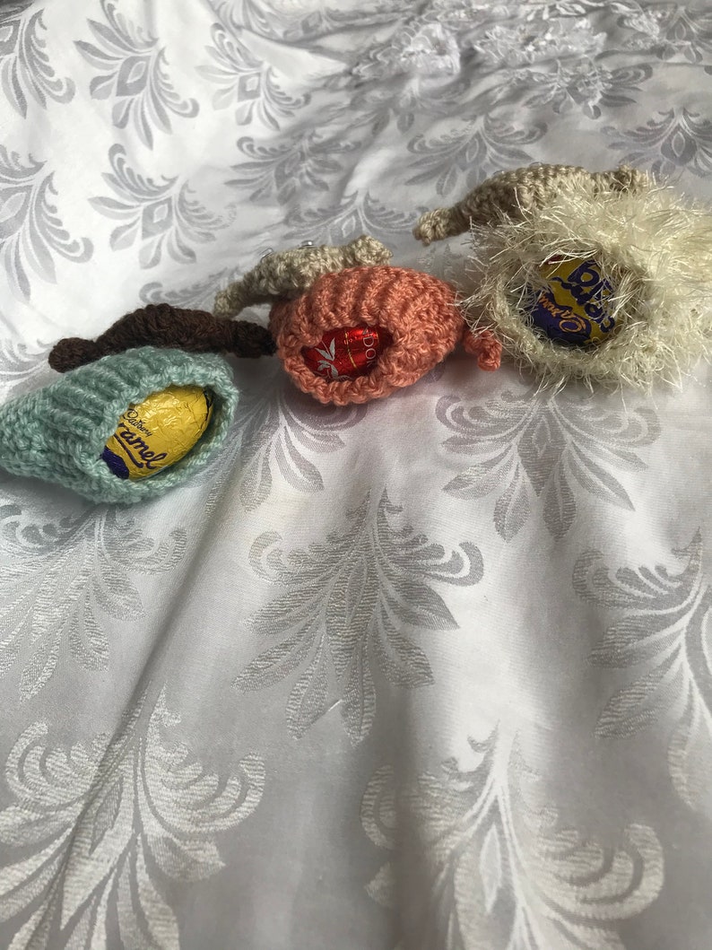 May include: Three crocheted Easter egg covers, one in light blue, one in pink, and one in white. Each cover has a chocolate egg inside, with the brand names "Cadbury Caramel", "Cadbury Dairy Milk", and "Cadbury Creme Egg" visible.