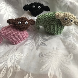 May include: Three knitted sheep toys, one black, one green, and one pink. The sheep have black eyes and white faces.