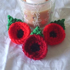 May include: Three handmade crochet poppies in vibrant red with black centers and green leaves. The flowers are arranged on a white surface, with a glass candle holder in the background.