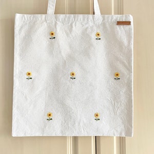 May include: White canvas tote bag with embroidered yellow sunflowers. The bag has a brown leather tag with the text "The New Normal" on it.