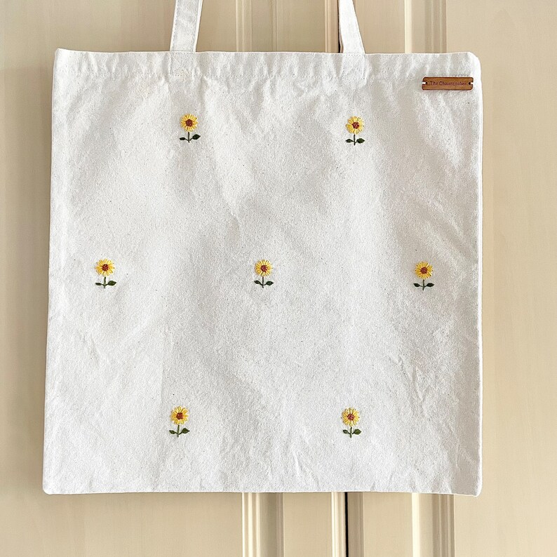 May include: White canvas tote bag with embroidered yellow sunflowers. The bag has a brown leather tag with the text "The New Normal" on it.