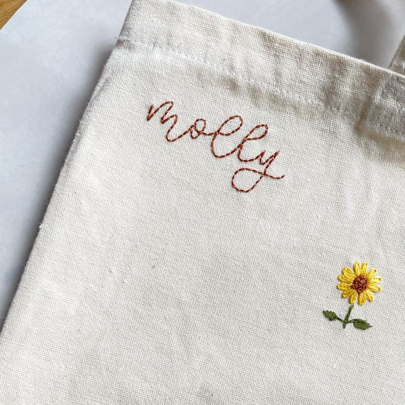 May include: A white canvas tote bag with the name "molly" embroidered in brown thread. A small yellow sunflower is embroidered in the bottom right corner.