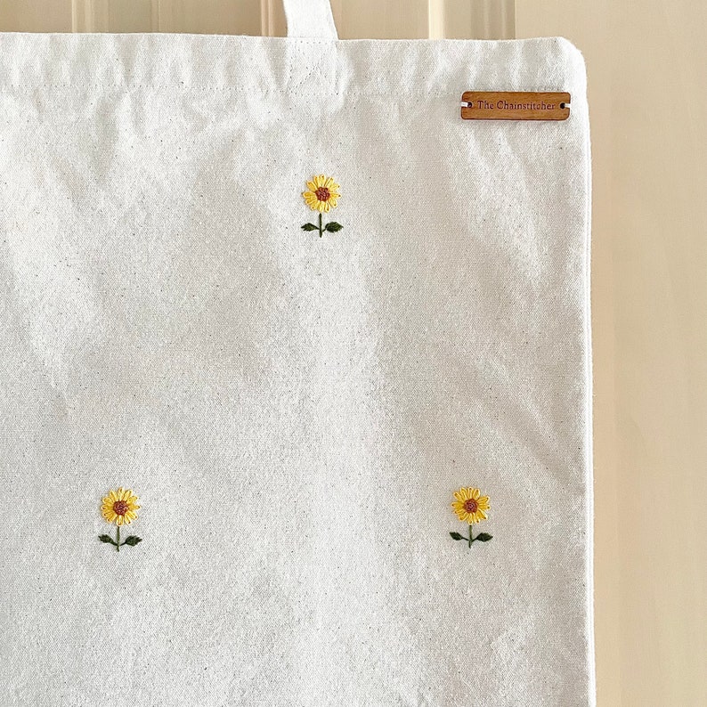 May include: White canvas tote bag with three embroidered yellow sunflowers. The bag has a brown leather tag with the text "The Chainstrucker" printed on it.