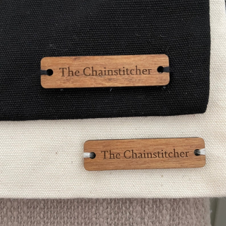 May include: Two wooden tags with the text "The Chainstitcher" on them. The top tag is on a black fabric background and the bottom tag is on a white fabric background.
