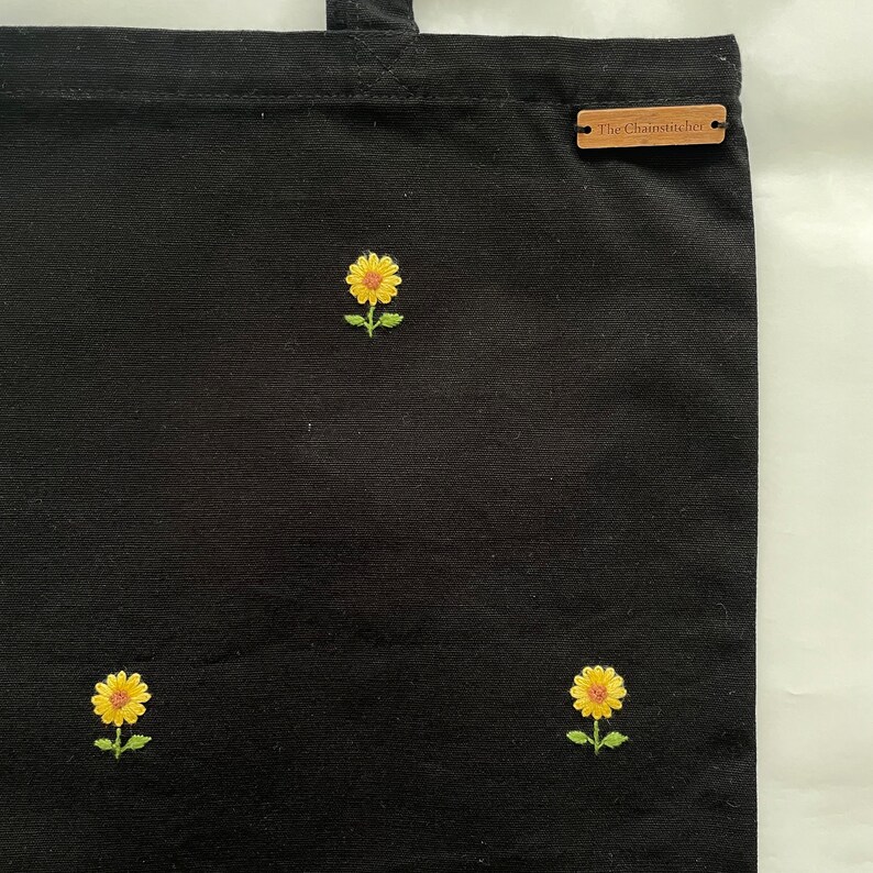 May include: Black canvas tote bag with three embroidered yellow sunflowers. The bag has a brown leather tag with the text "The Chainstitcher" on it.