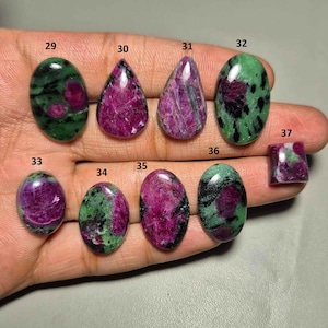 Puede incluir: Una colección de gemas pulidas, principalmente rubí en zoisita, mostradas en una mano. Las piedras varían en forma, incluyendo ovaladas, en forma de lágrima y cuadradas. Las secciones de rubí son de un rojo intenso, contrastando con la zoisita verde y negra.
