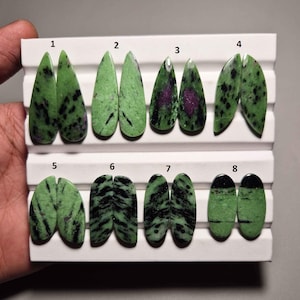 May include: A collection of eight pairs of polished zoisite gemstones, each with a unique pattern. The stones are primarily green with black and purple inclusions. They are displayed on a white tray, showcasing their varied shapes and colors.
