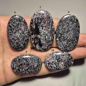 Może przedstawiać: Pięć owalnych kamieni szlachetnych o nakrapianym wyglądzie. Kamienie mają ciemny kolor bazowy z mieszanką białych i czerwonofioletowych plam. Kamienie szlachetne są trzymane w dłoni, prezentując ich wypolerowane powierzchnie.