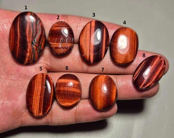 May include: Eight polished oval-shaped red tiger eye gemstones. The stones display a range of rich red and brown hues with visible chatoyancy. Each stone is unique, showcasing natural patterns and variations in color and texture. The gemstones are displayed on a neutral background.