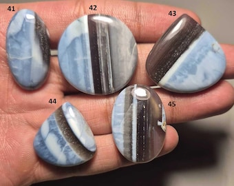 May include: Five polished opal gemstones in various shapes: oval, round, and triangular. The opals feature a mix of blue, brown, and white colors, with unique patterns and striations. Each stone is numbered for identification.