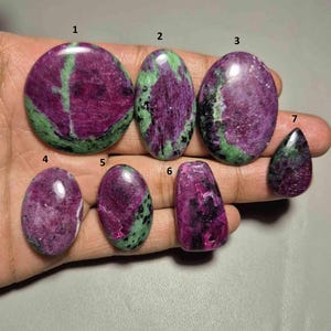 May include: Seven polished ruby zoisite gemstones in various shapes. The stones display a mix of deep magenta, green, and black colors. The gemstones are oval, round, and teardrop-shaped, showcasing natural patterns and textures.