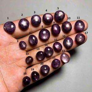 May include: A collection of twenty-two polished, round and oval cabochon gemstones in shades of deep purple and plum. Each stone displays a distinct star-like light reflection on its surface, and each is labeled with a small black number from 1 to 22.