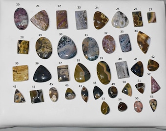 May include: A collection of polished gemstones in various shapes and colors, including oval, rectangular, and teardrop forms. The stones display a range of hues, such as brown, gray, pink, and yellow. Each stone is numbered for identification.