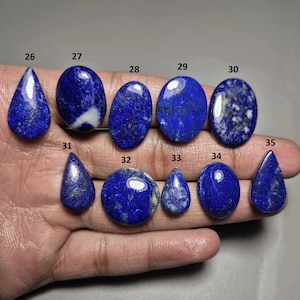 Puede incluir: Una colección de piedras preciosas de lapislázuli pulidas en varias formas, incluyendo ovaladas, en forma de lágrima y redondas. Las piedras azul oscuro tienen motas doradas y vetas blancas. Las piedras preciosas se muestran en una mano.