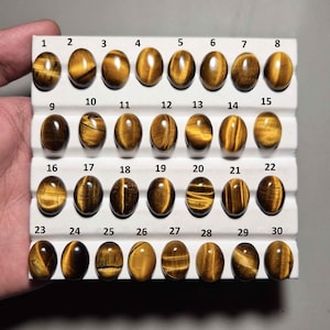 May include: A collection of thirty oval-shaped tiger's eye gemstones, each with unique patterns of golden brown and dark brown hues. The gemstones are arranged in rows on a white display card, numbered from 1 to 30.