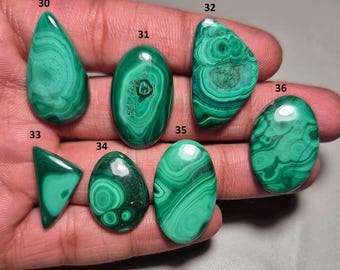 Puede incluir: Una colección de piedras preciosas de malaquita pulidas en varias formas, incluyendo ovaladas, en forma de lágrima y triangulares. Las piedras muestran un color verde vibrante con patrones arremolinados distintivos. Las piedras preciosas se sostienen en una mano.