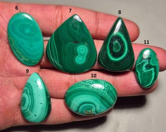 Puede incluir: Una colección de piedras preciosas de malaquita pulidas en varias formas, incluyendo ovaladas, de lágrima y redondeadas. Las piedras muestran un rico patrón arremolinado de tonos verdes oscuros y claros, con un acabado brillante. Las piedras preciosas se sostienen en una mano.