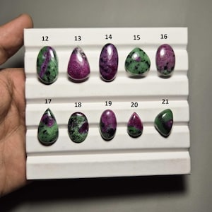 May include: A selection of polished ruby zoisite gemstones in diverse shapes, such as oval, teardrop, and triangular. The stones showcase a blend of deep pink and green colours, presented on a white display tray.